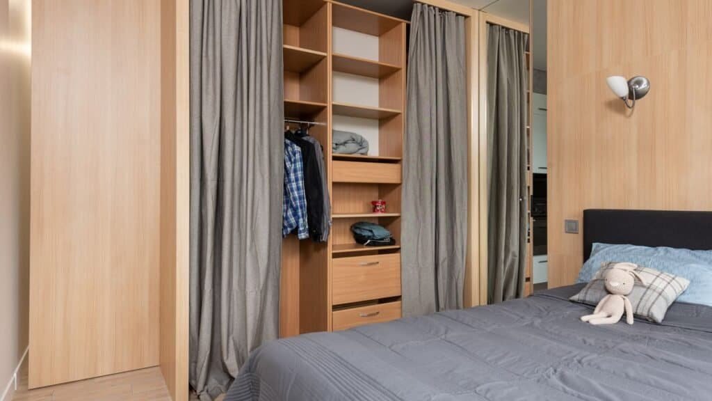 Renter-friendly storage in a small apartment bedroom: an open wardrobe with hanging clothes and folded items on shelves beside a made bed