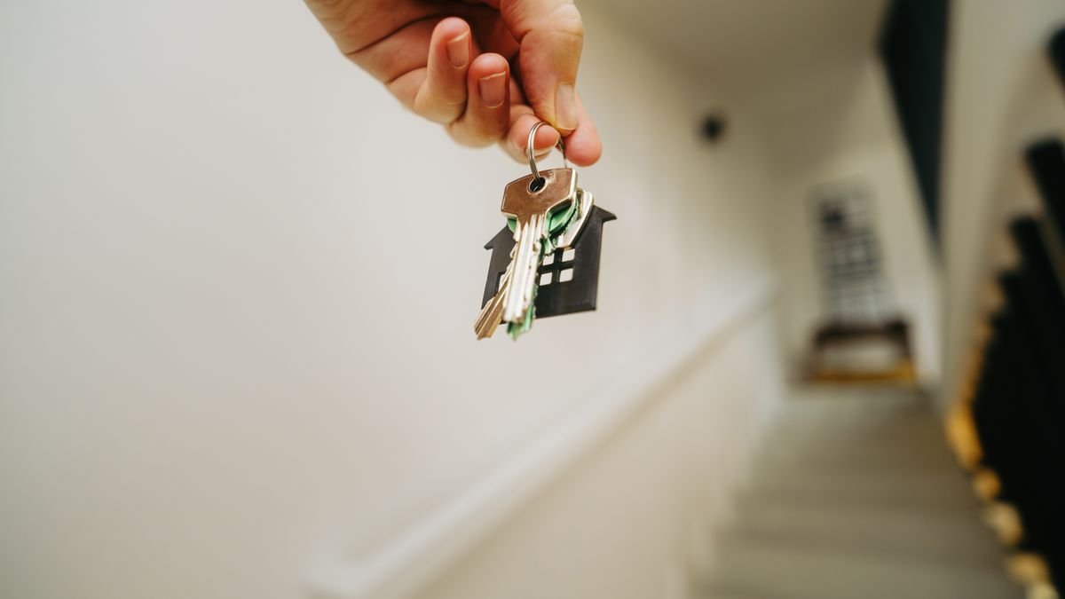A hand holding house-shaped keys outside an apartment hallway, representing the move-in point when renters insurance should already be in place