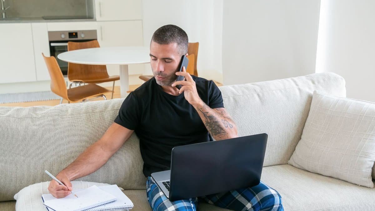 young renter on a phone call taking notes with a laptop nearby, daylight apartment