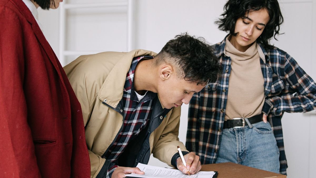 three renters signing paperwork on a moving box in an empty apartment